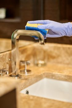 Close up of maid hand in a protective glove holding a washcloth and cleaning the tap. Hotel service concept