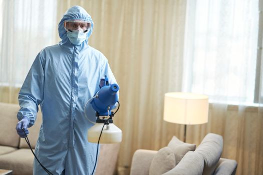 Man in protective suit and glasses using microparticle gun for virus prevention in apartments. Coronavirus and quarantine concept