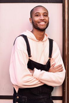 Young black man wearing casual clothes smiling against pink urban background. Attractive african guy with bib pants outdoors