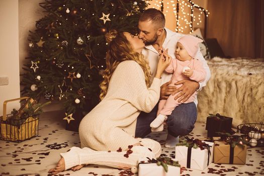 Portrait of kissing parents holding baby daughter playing with confetti in father’s arms. Christmas.