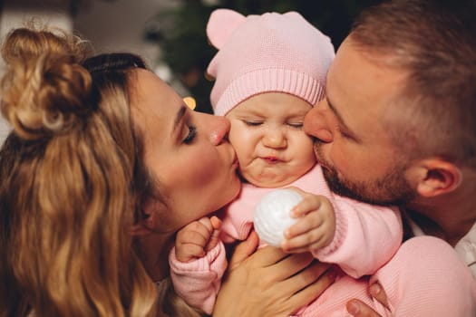 Close-up portrait of loving parents kissing their cute baby daughter in pink clothing at xmas.