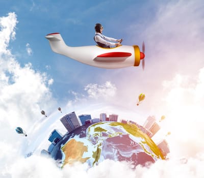 Cheerful young pilot sitting in cabin of small airplane. Funny man in aviator hat with goggles driving propeller plane. Traveling around the world by airplane. Earth globe among cloudy skyscapes.