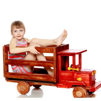 Beautiful little caucasian girl with a short bangs and hair tucked into a tail. In striped in a T-shirt and shorts.The girl is sitting on a large toy wooden car.