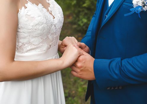 The groom in a blue suit holds the bride's hands in a white dress. Tenderness and affection at the wedding.
