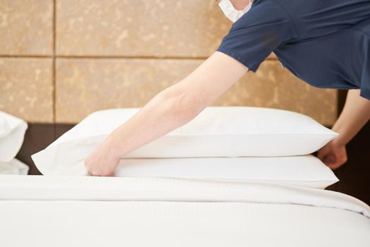 Cropped of maid in mask adjusting white pillows on the bed in hotel room. Hotel service concept