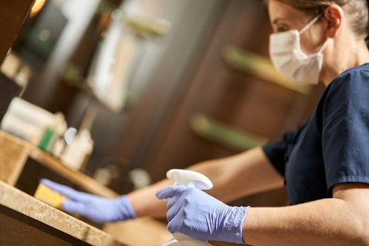 Side view of maid in mask holding spray and washcloths and cleaning in hotel bathroom. Hotel service concept