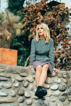 Happy young blond woman sitting on urban autumn background. Smiling blonde girl with flowered dress enjoying life outdoors.