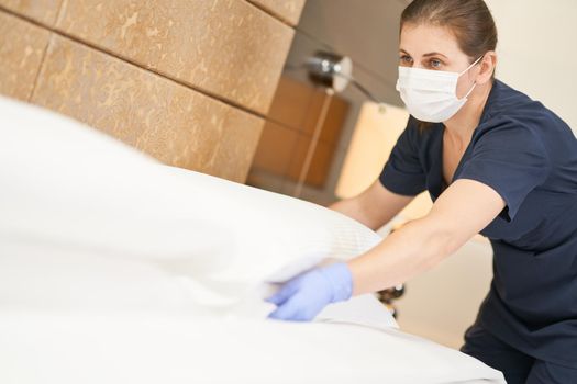 Maid in mask fluffing white pillow on bed while cleaning the room in hotel. Hotel service concept