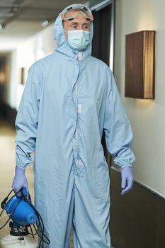 Male specialist in hazmat suit with disinfectant in hotel while walking in corridor. Coronavirus and quarantine concept