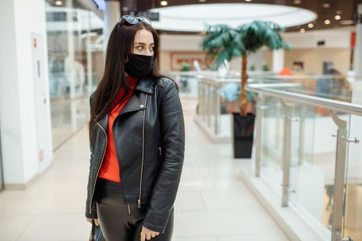 A girl with a medical black mask is walking along a shopping center. Coronavirus pandemic. A woman with a mask is standing in a shopping center. A girl in a protective mask is shopping at the mall.
