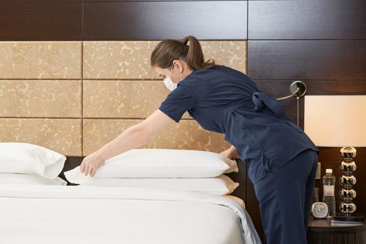 Side view of maid in mask making bed in hotel room while holding pilows. Hotel service concept