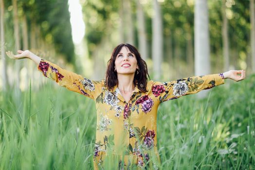 Enjoying the nature. Young woman arms raised enjoying the fresh air in green forest. Female wearing flowered dress.