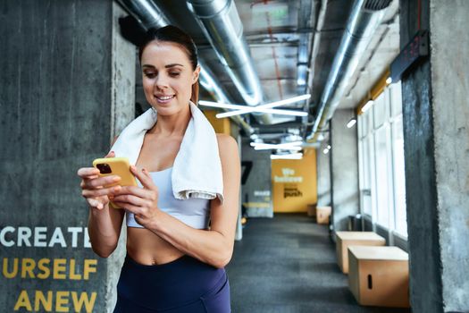 Chatting with friend. Young focused fitness woman in sportswear using her smartphone, texting sms while standing at gym, resting after workout. Sport, wellness and healthy lifestyle