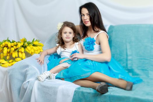 Beautiful young mother and little daughter in the same blue dresses on the sofa with a bouquet of flowers.