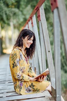 Young woman, wearing flowered dress, reading a book on a rural bridge.