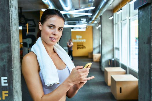 Young beautiful fitness woman in sportswear holding her smartphone and looking at camera while standing at gym after workout. Sport, wellness and healthy lifestyle