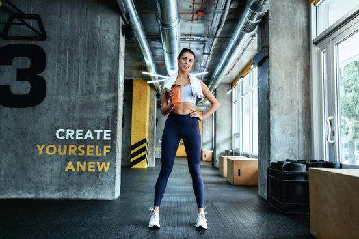 Full length of a young beautiful fitness woman in sportswear holding bottle of water and smiling at camera while standing at industrial gym, resting after workout. Sport, wellness and healthy lifestyle