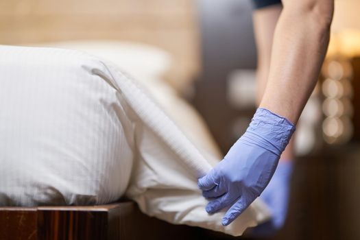 Cropped photo of maid in gloves making regular bed linen change in hotel room. Hotel service concept