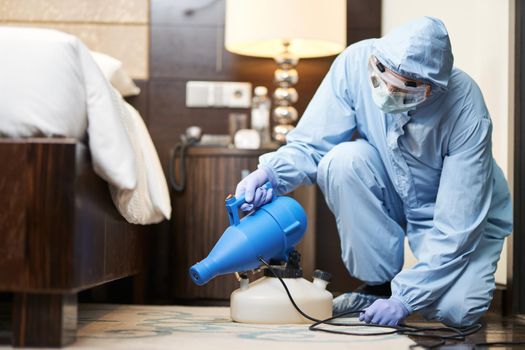 Man in biohazard suit and gloves disinfecting bedroom in hotel. Coronavirus and quarantine concept