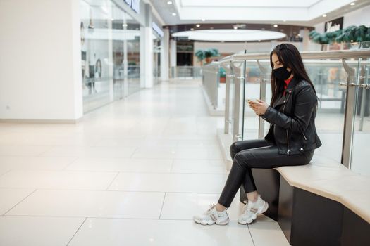 Girl with medical black mask and mobile phone in a shopping center. Coronavirus pandemic. A woman with a mask is standing in a shopping center. A girl in a protective mask is shopping at the mall.