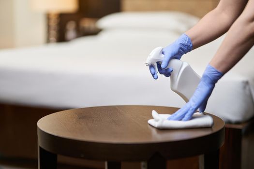 Cropped photo of maid spraying disinfectant on furniture while cleaning the room. Housekeeping and hygiene concept