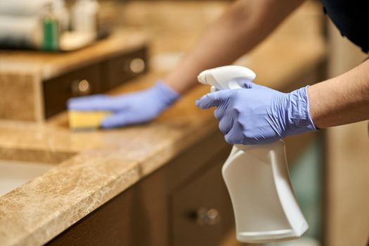 Close up of chambermaid in gloves splashing disinfectant on the bathroom surface in hotel room. Hotel service concept