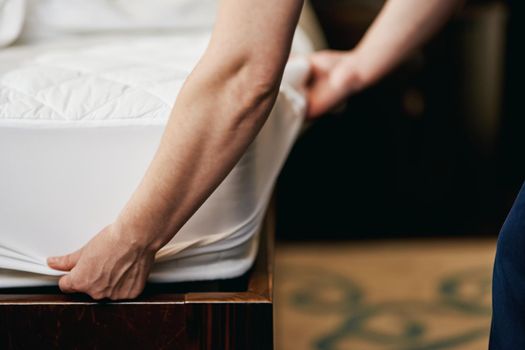 Cropped photo of chambermaid changing clean sheet on white mattress in hotel room. Hotel service concept