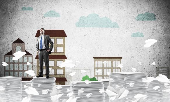 Confident businessman in suit standing on pile of documents among flying paper planes with drawn cityscape background. Mixed media.