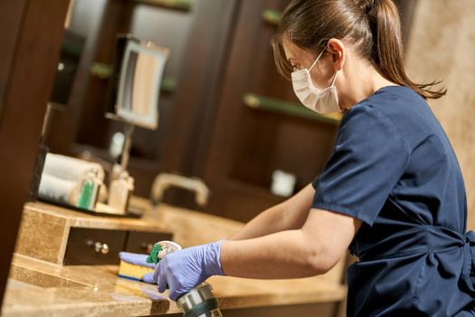 Side view of maid in mask splashing disinfectant on the bathroom surface. Hotel service concept