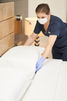 Female housemaid in protective mask fluffing white pillow on bed in modern hotel room. Hotel service concept