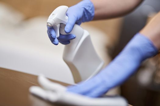 Close up of of housemaid in gloves spraying disinfectant on furniture while cleaning hotel room. Housekeeping and hygiene concept