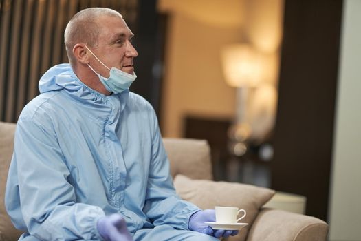 Side view of worker in protective suit sitting on sofa while drinking coffee in room. Coronavirus and quarantine concept