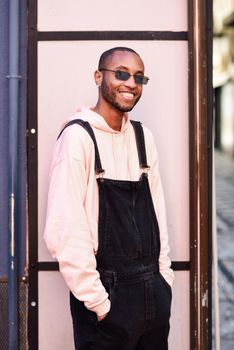 Young black man wearing casual clothes and sunglasses, smiling against pink urban background. Happy Young African guy with bib pants outdoors