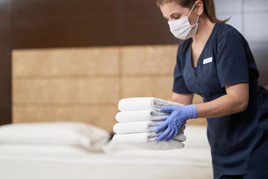 Cropped photo of chambermaid in mask putting fresh towels on bed in hotel room. Copy space. Hotel service concept