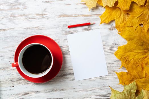 Flat lay autumn composition with cup of black tea and yellow autumn leaves. Time of tea break concept. Hot drink and blank paper sheet with pen lies on vintage wooden desk with bright foliage.