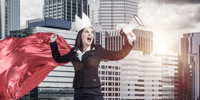Young confident businesswoman wearing red cape against modern city background screaming in megaphone