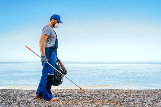 Man volunteer in uniforn collecting garbage on the beach with a reach extender stick