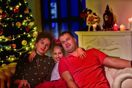 A happy family, mom, dad and little daughter, near a beautifully decorated Christmas tree with toys and bright lights, near a large panoramic window. New Year concept, family vacation.