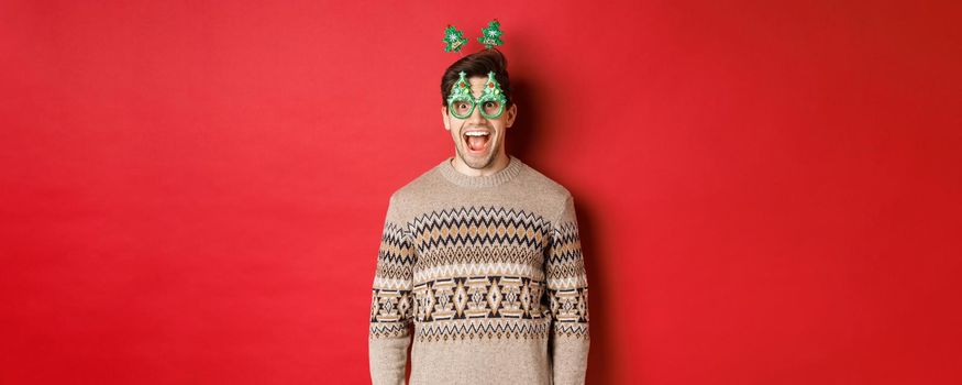 Portrait of surprised and excited handsome guy in party glasses and winter sweater, celebrating christmas and having fun, standing against red background.