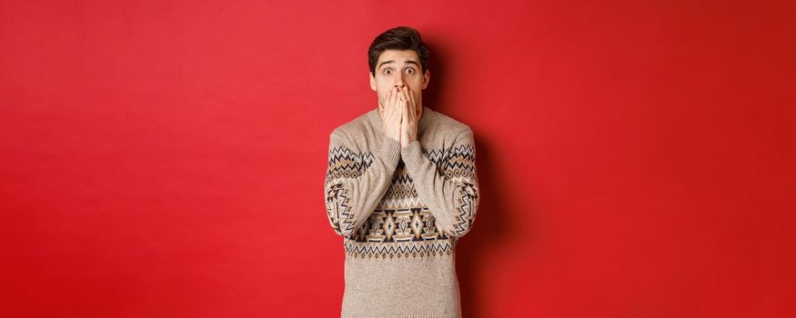 Portrait of shocked and speechless caucasian guy in christmas sweater, reacting to announcement, standing startled against red background.