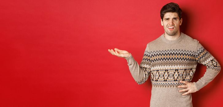 Image of confused and disappointed handsome man, complaining about christmas party, raising hand and looking bothered, standing over red background.