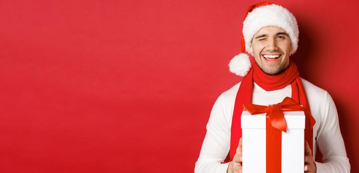 Concept of winter holidays, christmas and lifestyle. Handsome cheeky man in santa hat and scarf, holding present and smiling, winking at camera, standing over red background.