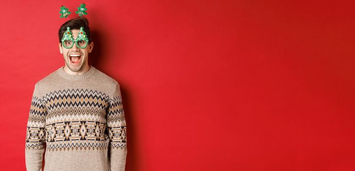 Portrait of surprised and excited handsome guy in party glasses and winter sweater, celebrating christmas and having fun, standing against red background.