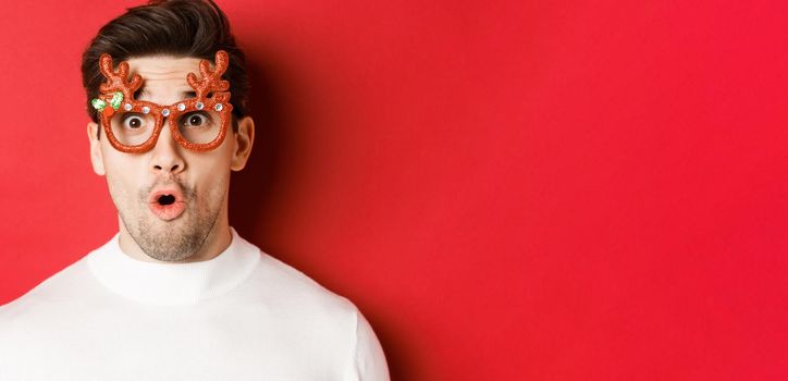 Concept of winter holidays, christmas and celebration. Close-up of attractive guy with bristle, wearing new year party glasses and looking impressed at promo offer, standing over red background.