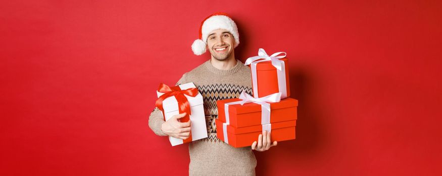 Concept of winter holidays, new year and celebration. Portrait of lovely smiling man prepared gifts for christmas, holding presents and looking at camera heartwarming, red background.