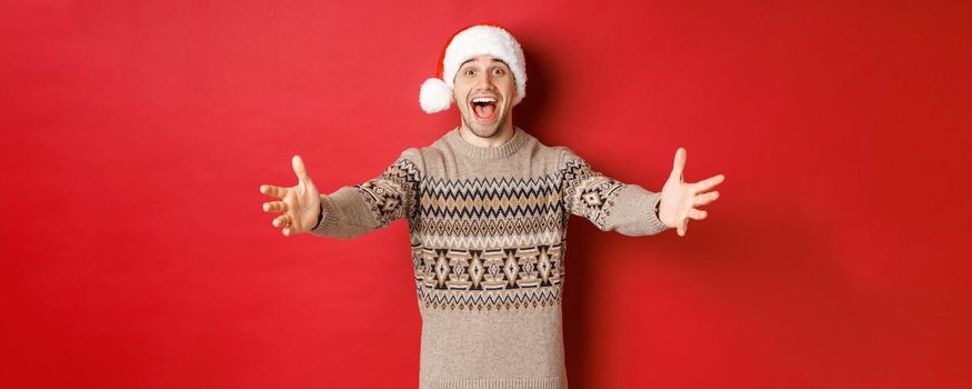 Portrait of happy, attractive man in christmas sweater and santa hat, reaching hands for gift, want to take something and smiling, standing over red background.