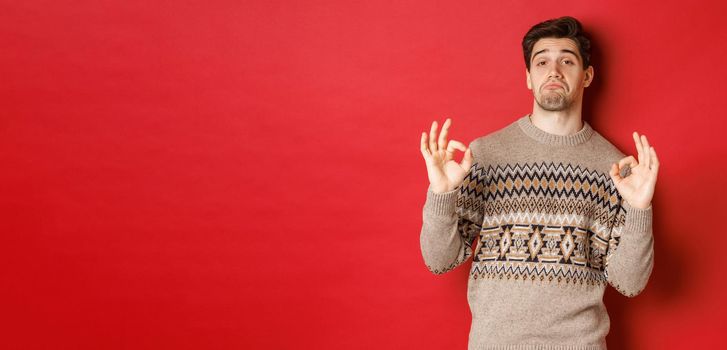 Impressed handsome man in christmas sweater, showing okay signs and praising good party, nodding in approval, standing pleased against red background.