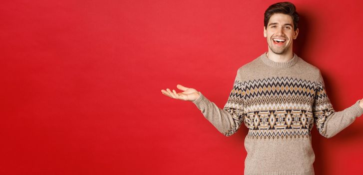 Portrait of happy good-looking man celebrating new year holidays, wearing christmas sweater, spread hands sideways and smiling, holding something on copy space, standing over red background.