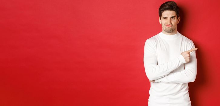 Doubtful and reluctant handsome man grimacing, pointing fingers right at something bad, standing in white sweater over red background.
