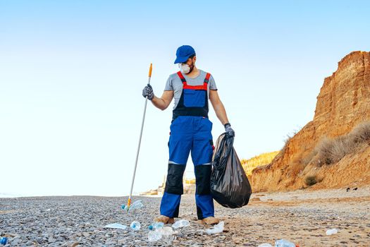 Man volunteer in uniforn collecting garbage on the beach with a reach extender stick